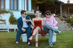 Three young adults in stylish attire sitting together on a bench outdoors, showing camaraderie and elegance.